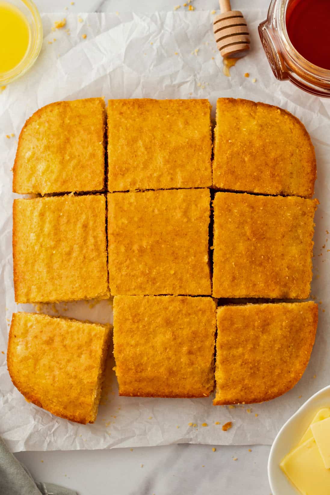 A square pan of cornbread cut into nine pieces, one piece slightly removed. Surrounding items include butter, honey, and a honey dipper.