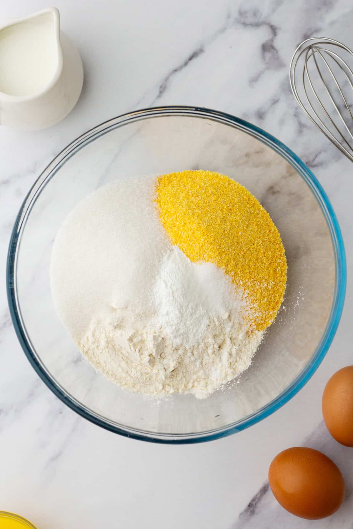 A glass bowl contains sugar, cornmeal, and flour, placed on a marble surface. Nearby are eggs, a whisk, and a small jug of milk.