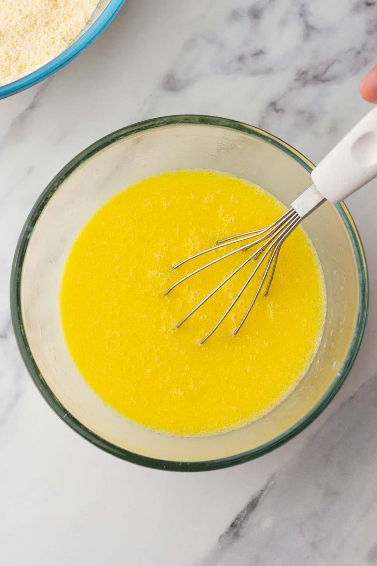 A whisk in a glass bowl with melted butter on a marble countertop, next to a bowl of dry ingredients.