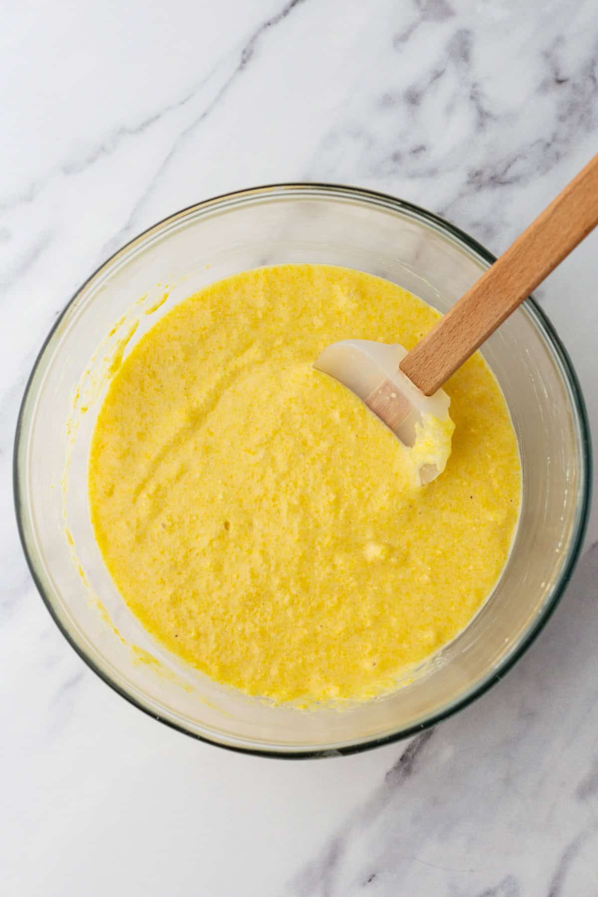 A glass bowl filled with yellow cornbread batter and a spatula on a marble countertop.