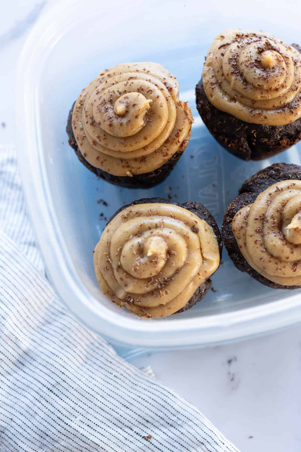 Four chocolate cupcakes with swirled buttercream frosting in a blue container, next to a striped cloth.