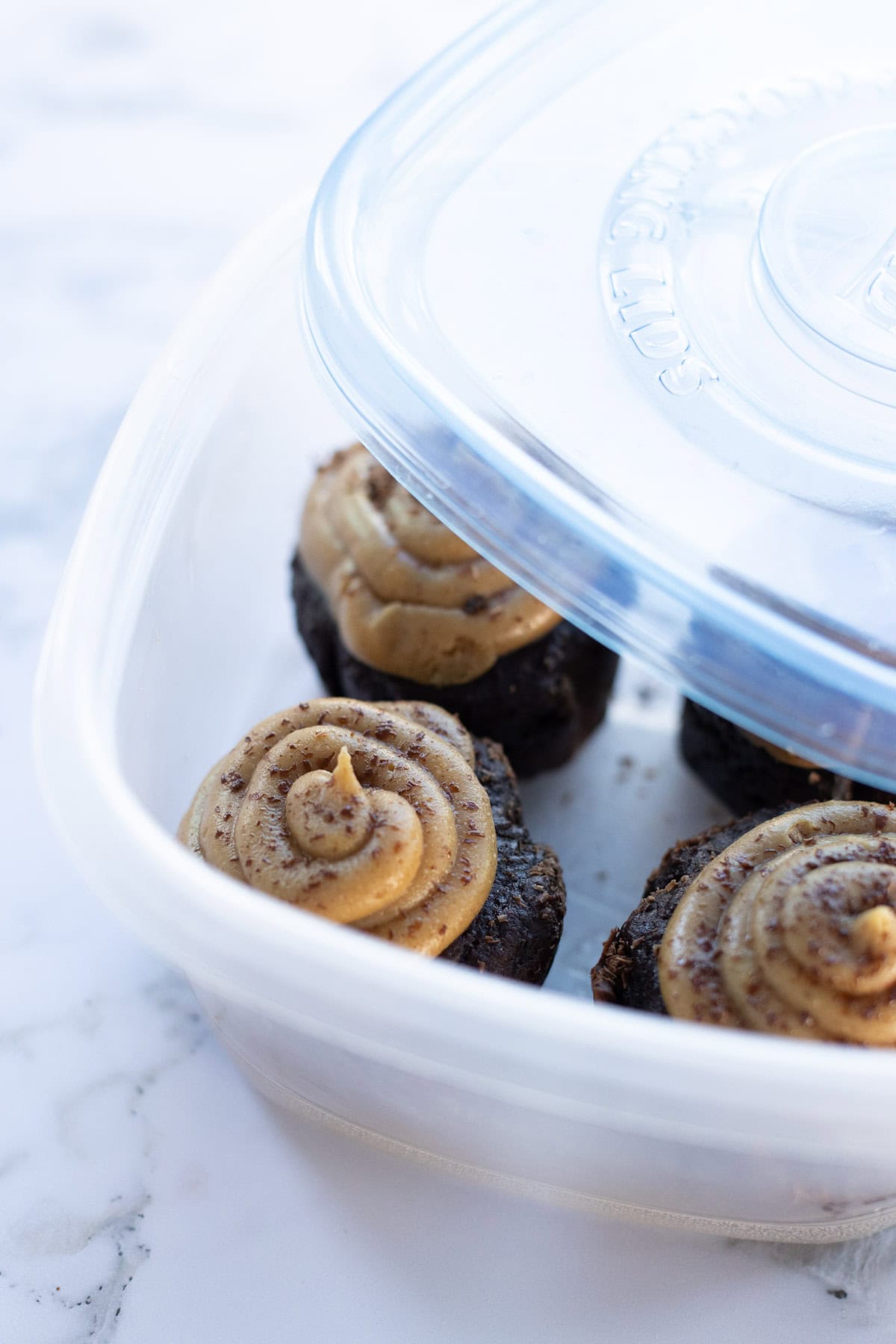 Four chocolate cupcakes with swirl Four chocolate cupcakes with swirled buttercream frosting in a plastic container with a partially closed lid on a marble surface.
