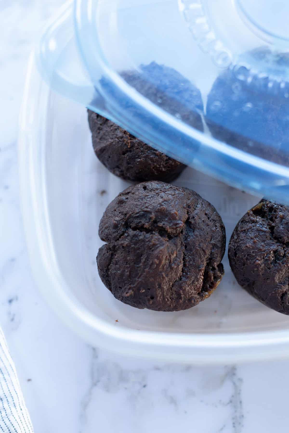 Three chocolate muffins in a transparent plastic container with a blue lid, placed on a marble surface.