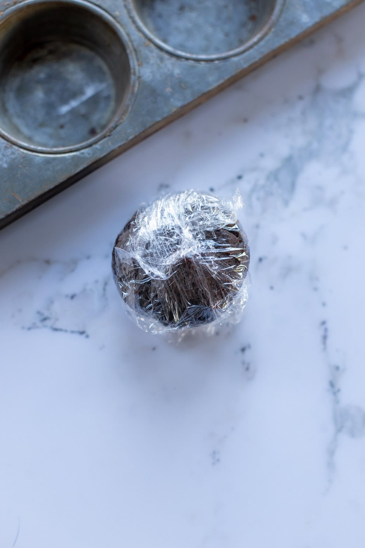Plastic-wrapped muffin on a marble surface next to a muffin tray.