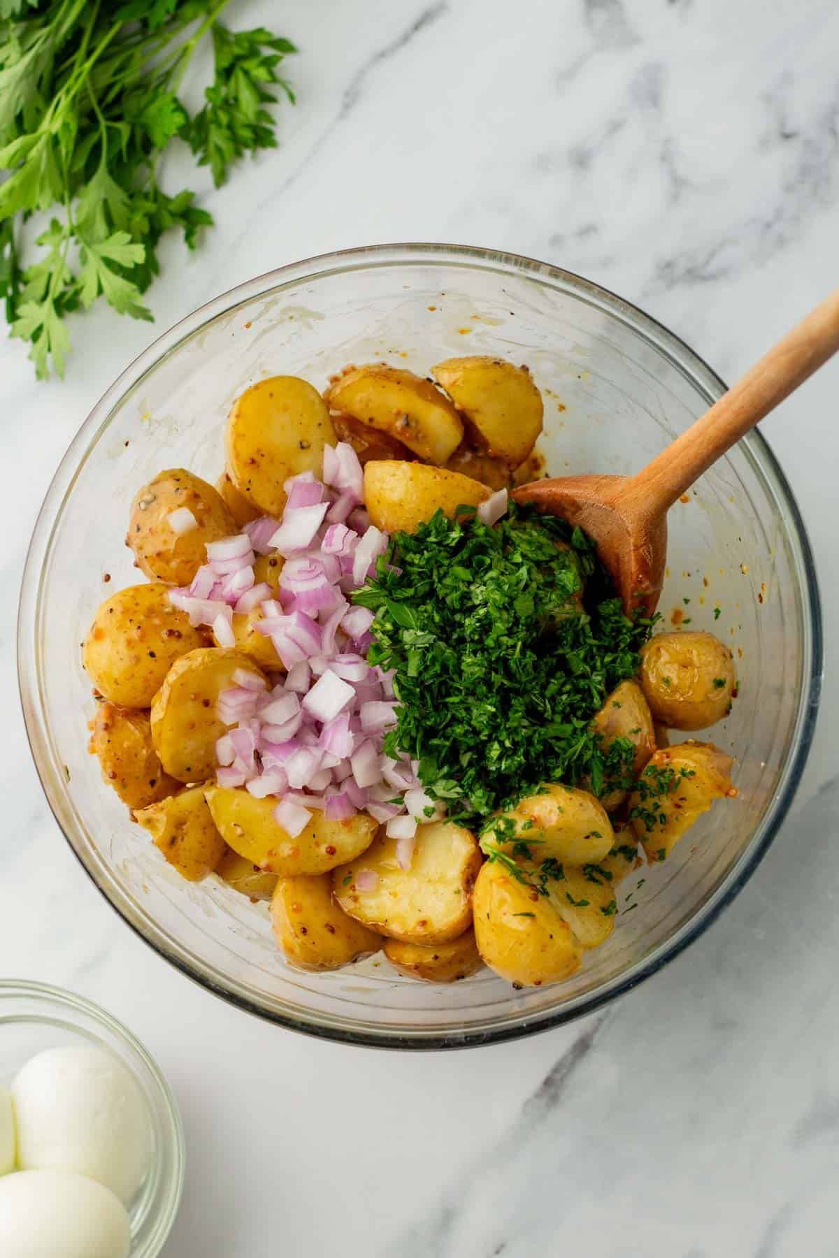 A bowl with seasoned potato salad ingredients: potatoes, chopped red onions, and herbs. A wooden spoon is in the bowl. Parsley and hard-boiled eggs are nearby on a marble surface.