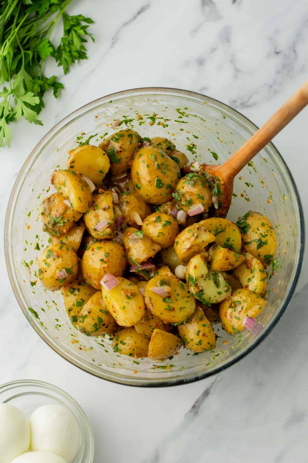 A bowl of potato salad with herbs and red onion on a marble surface, next to parsley and a bowl of eggs.