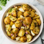 A bowl of potato salad with halved potatoes, chopped onions, and parsley on a marble surface. A plate with forks and parsley is in the background.