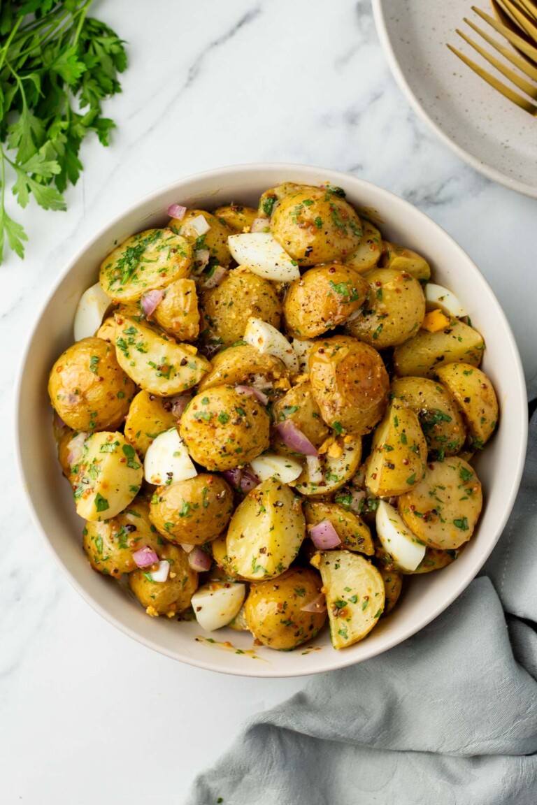 A bowl of potato salad with halved potatoes, chopped onions, and parsley on a marble surface. A plate with forks and parsley is in the background.