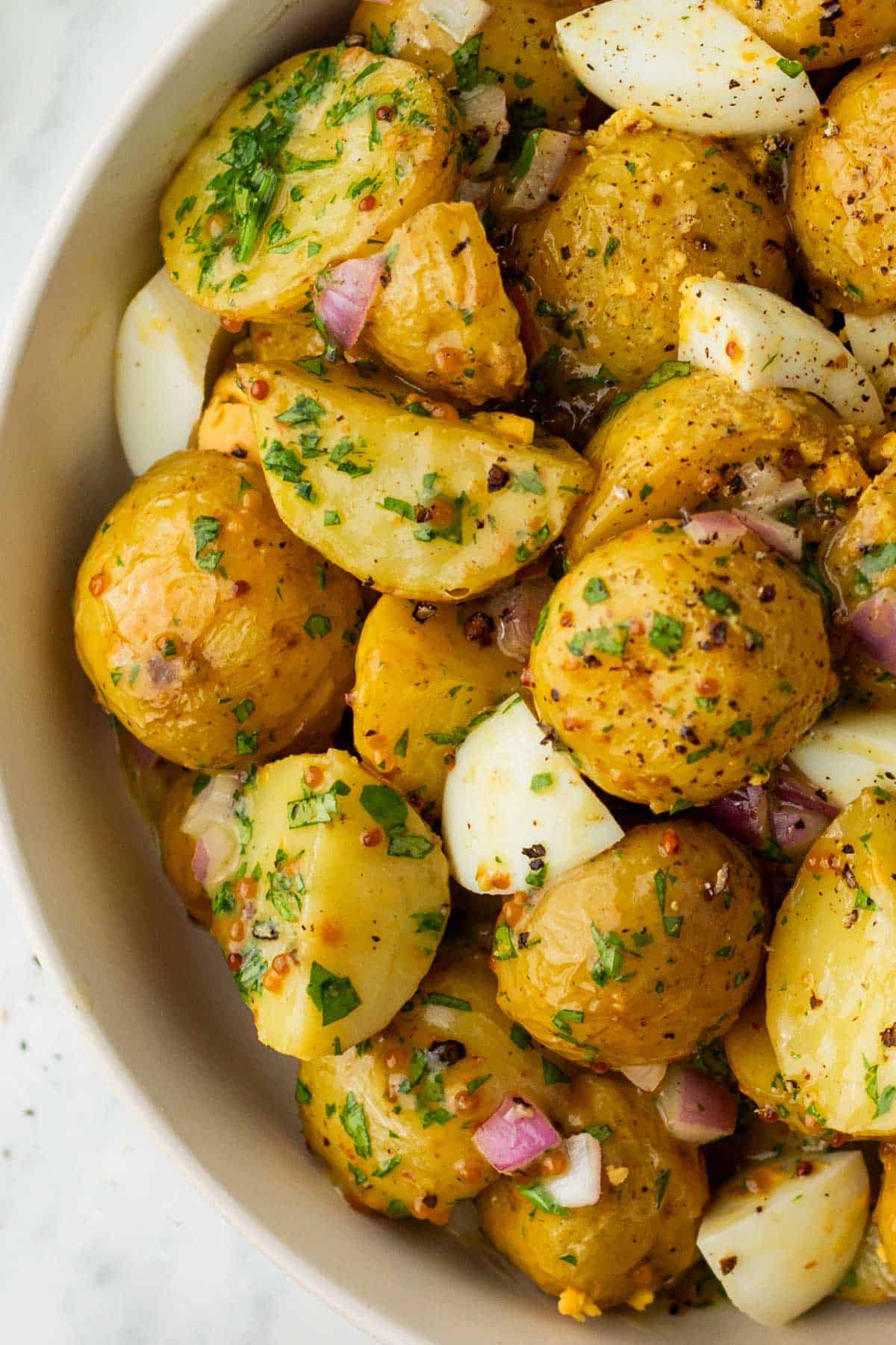 A bowl of potato salad with chopped potatoes, boiled eggs, red onions, and herbs, seasoned with black pepper and spices.