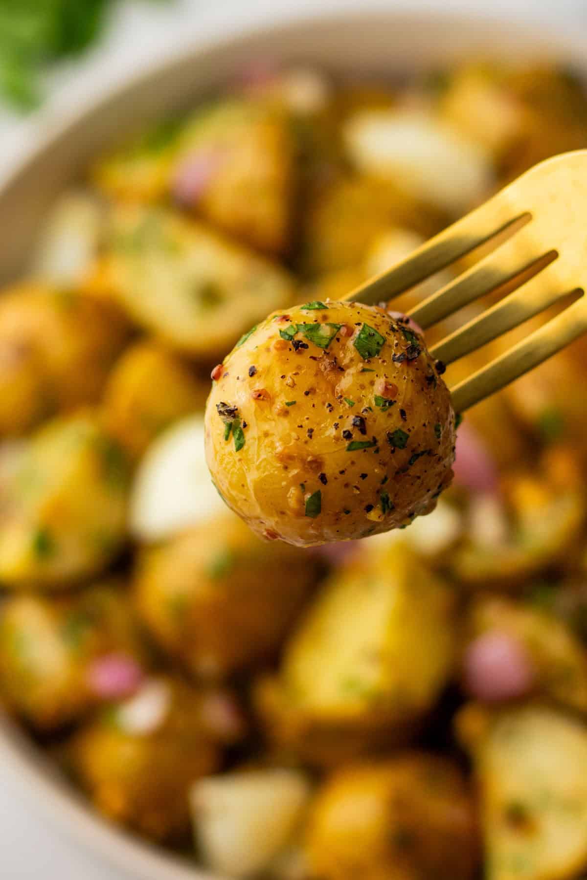 A fork holding a seasoned baby potato with herbs, against a blurred background of more seasoned potatoes in a bowl.