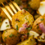 A fork holding a slice of seasoned potato with herbs and diced red onion. Surrounding are more potato pieces.