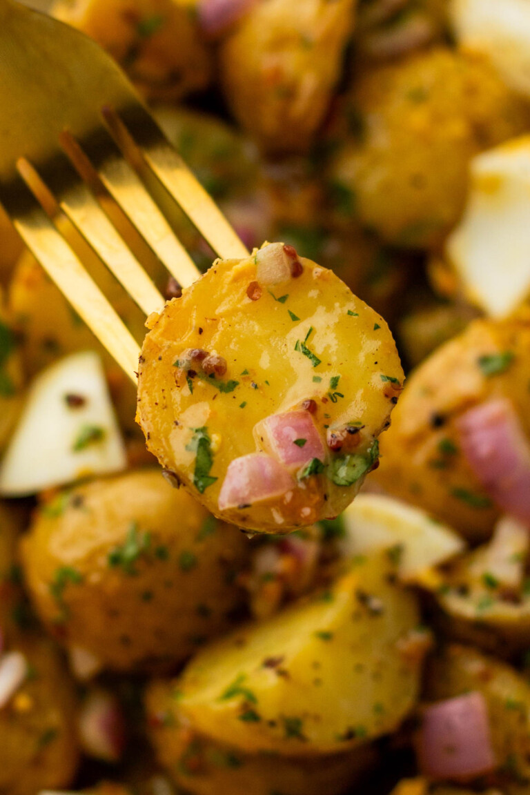 A fork holding a slice of seasoned potato with herbs and diced red onion. Surrounding are more potato pieces.