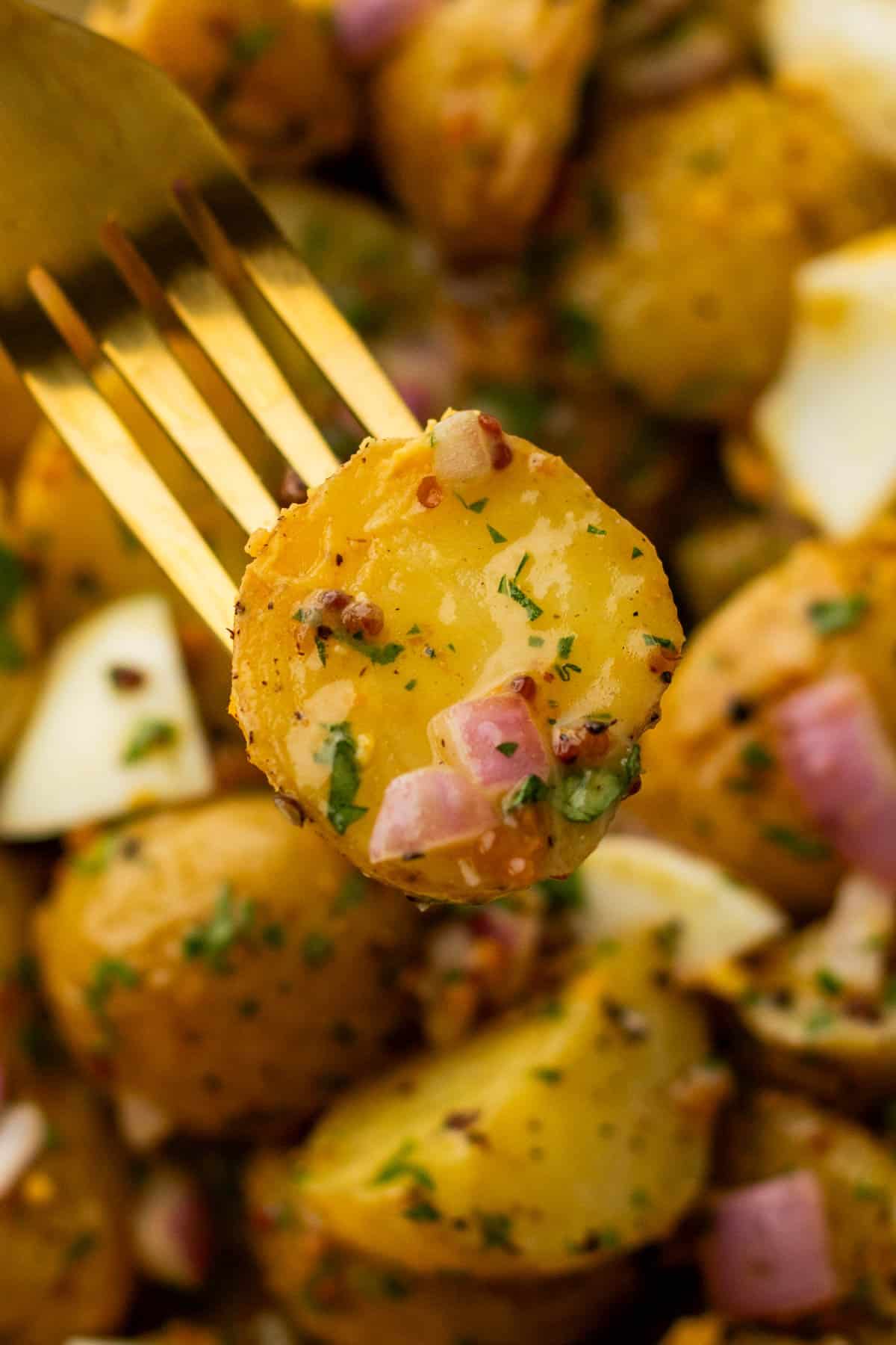 A fork holding a slice of seasoned potato with herbs and diced red onion. Surrounding are more potato pieces.