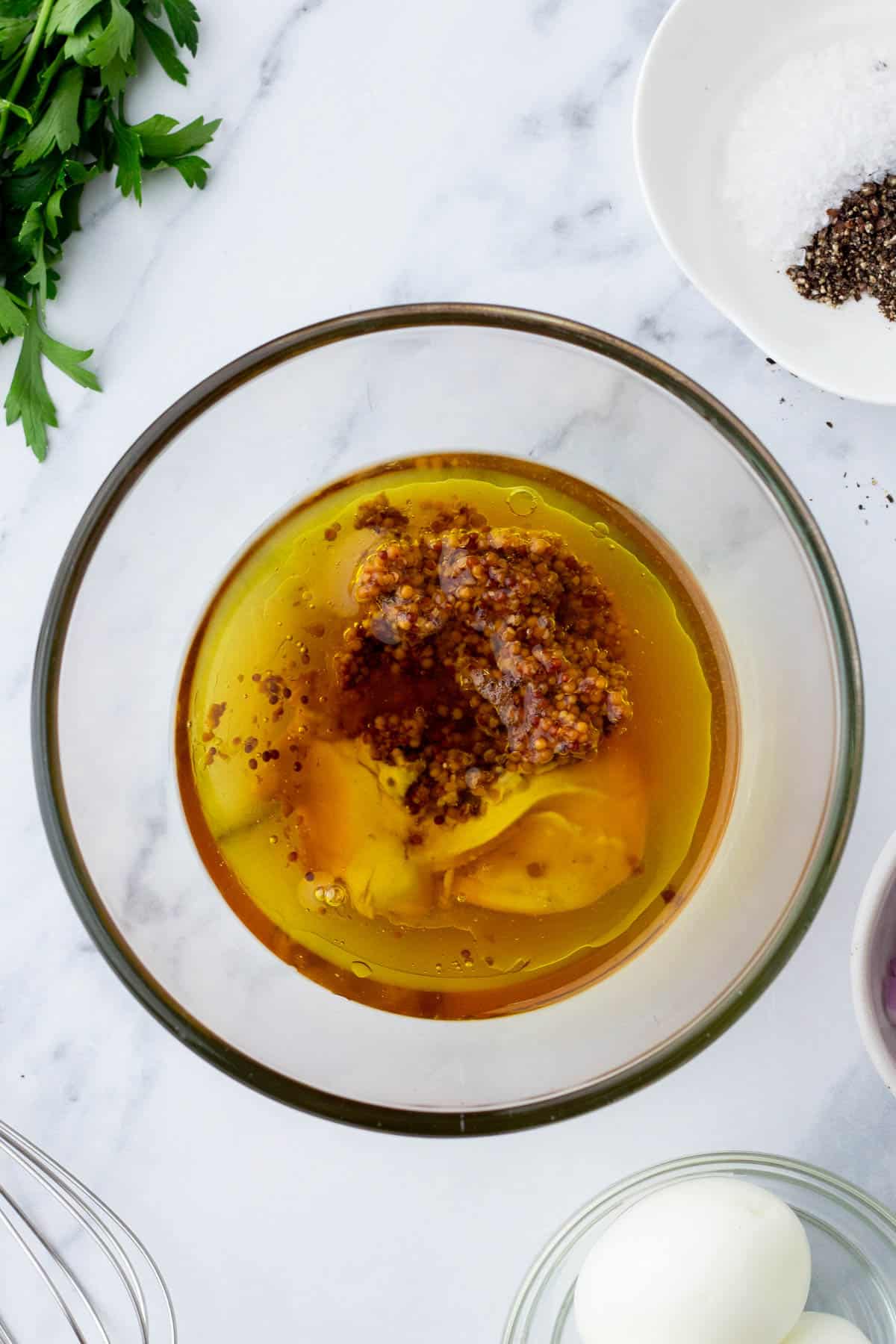 A bowl containing oil, mustard, and seasonings on a marble surface, surrounded by a whisk, parsley, a white plate with salt and pepper, and another bowl with an egg.