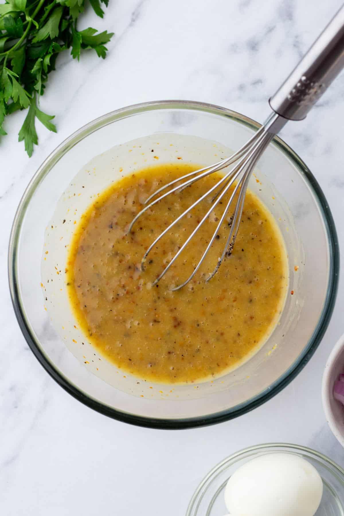 A glass bowl contains mustard vinaigrette with herbs, and a whisk. Nearby are parsley, peeled hard-boiled egg, and chopped onions.