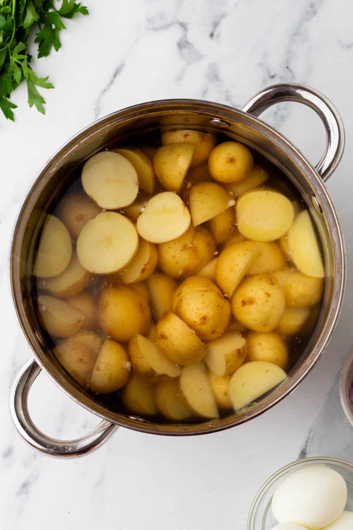 Pot filled with sliced potatoes submerged in water on a marble surface, next to a sprig of parsley.