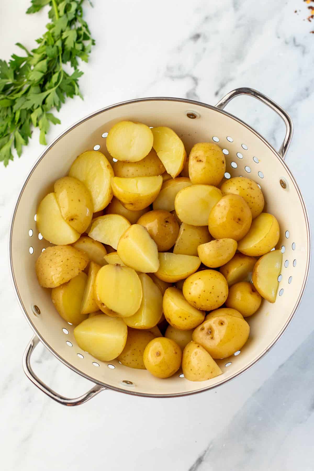 A colander filled with cut boiled yellow potatoes on a marble surface, with parsley on the side.
