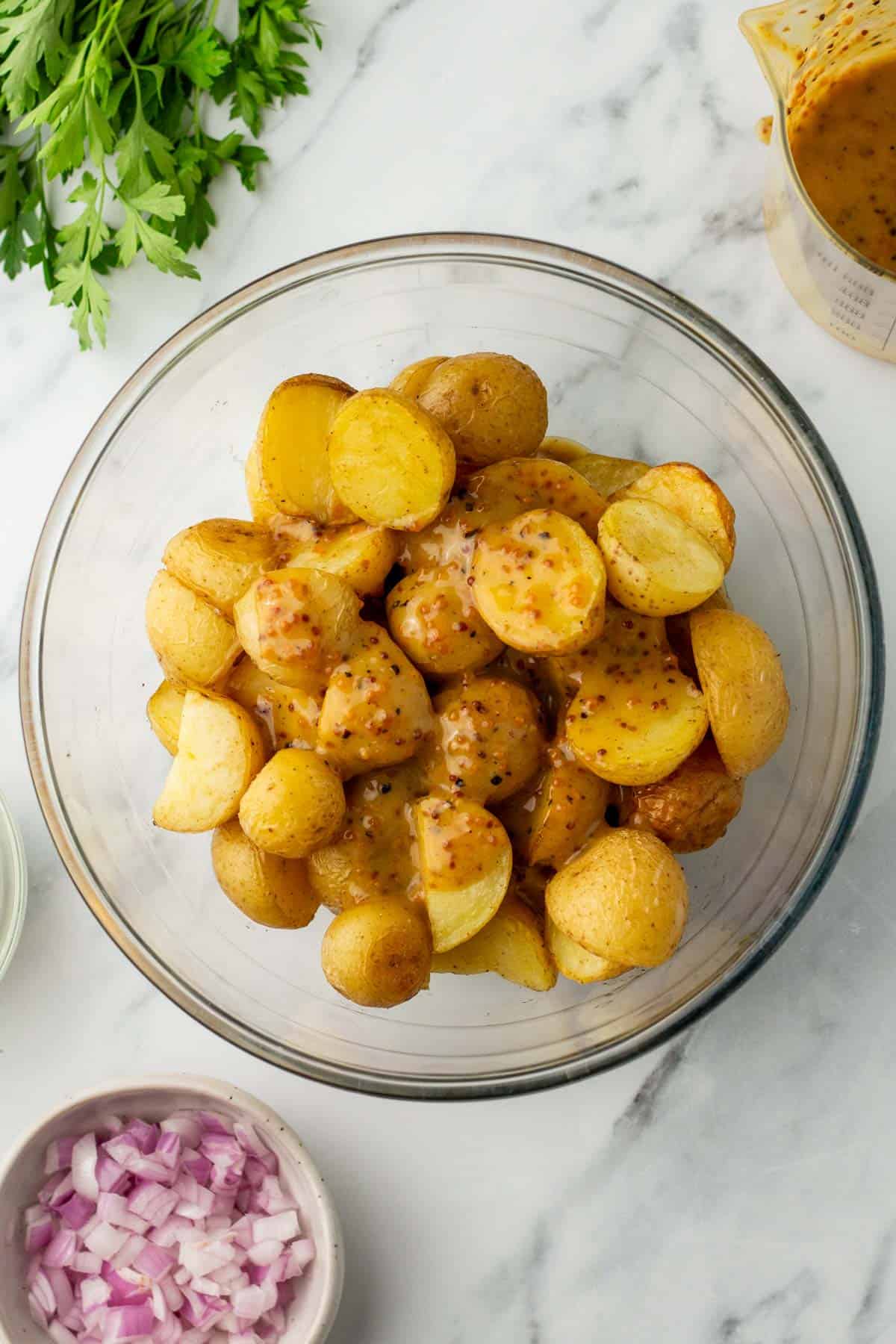 Bowl of yellow potatoes with mustard dressing next to chopped onions and parsley on a marble surface.