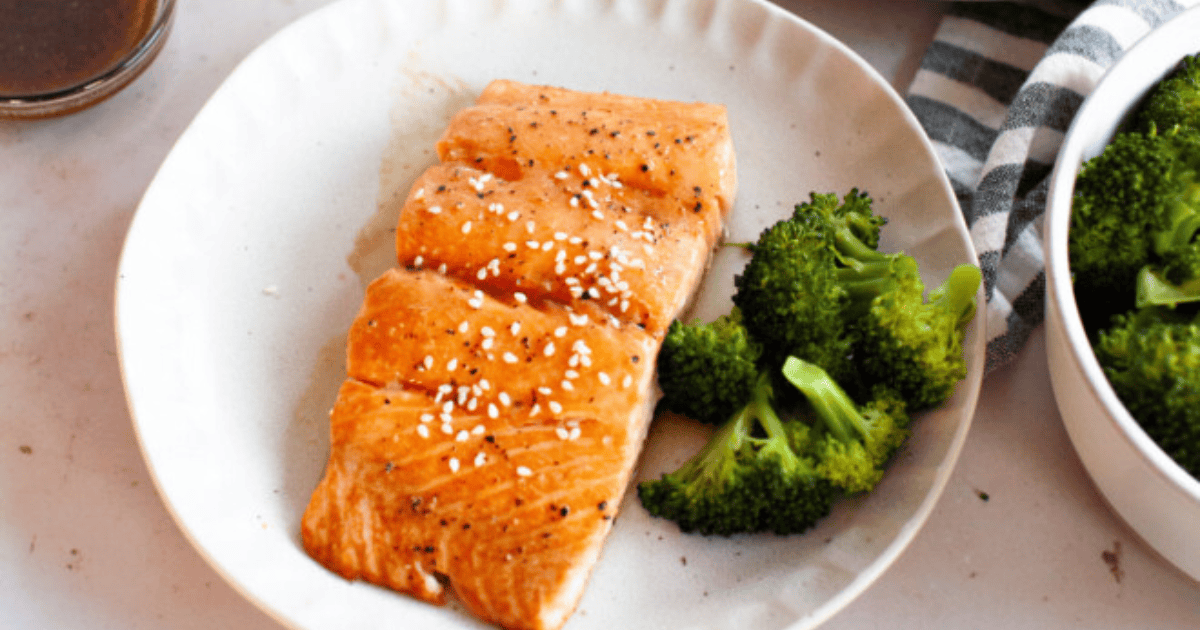 A plate with cooked salmon topped with sesame seeds and a side of broccoli. A striped cloth and bowl of broccoli are partially visible.