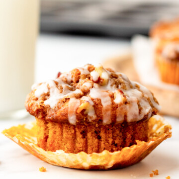 A muffin with white icing glaze sits in an unwrapped paper liner, placed on a marble surface. A blurred glass of milk is in the background.