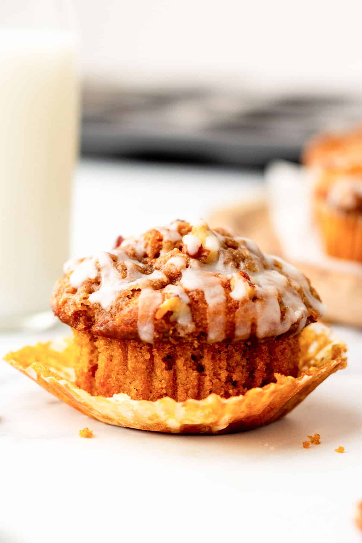 A muffin with white icing glaze sits in an unwrapped paper liner, placed on a marble surface. A blurred glass of milk is in the background.
