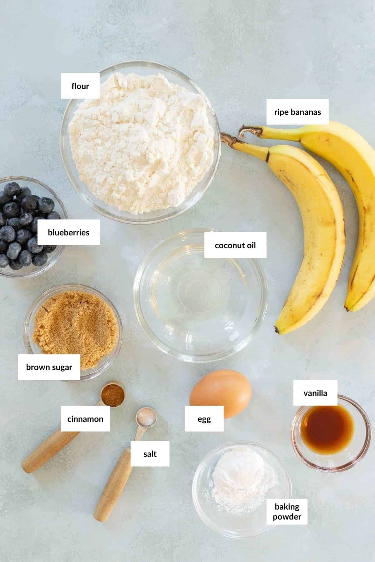 Ingredients for baking include flour, ripe bananas, blueberries, coconut oil, brown sugar, cinnamon, salt, an egg, vanilla extract, and baking powder, arranged on a light surface.
