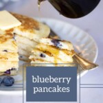 Golden syrup cascades over a tower of fluffy blueberry pancakes, each layer bursting with ripe berries. A fork sits invitingly beside the plate, ready to dive into this delectable morning treat.