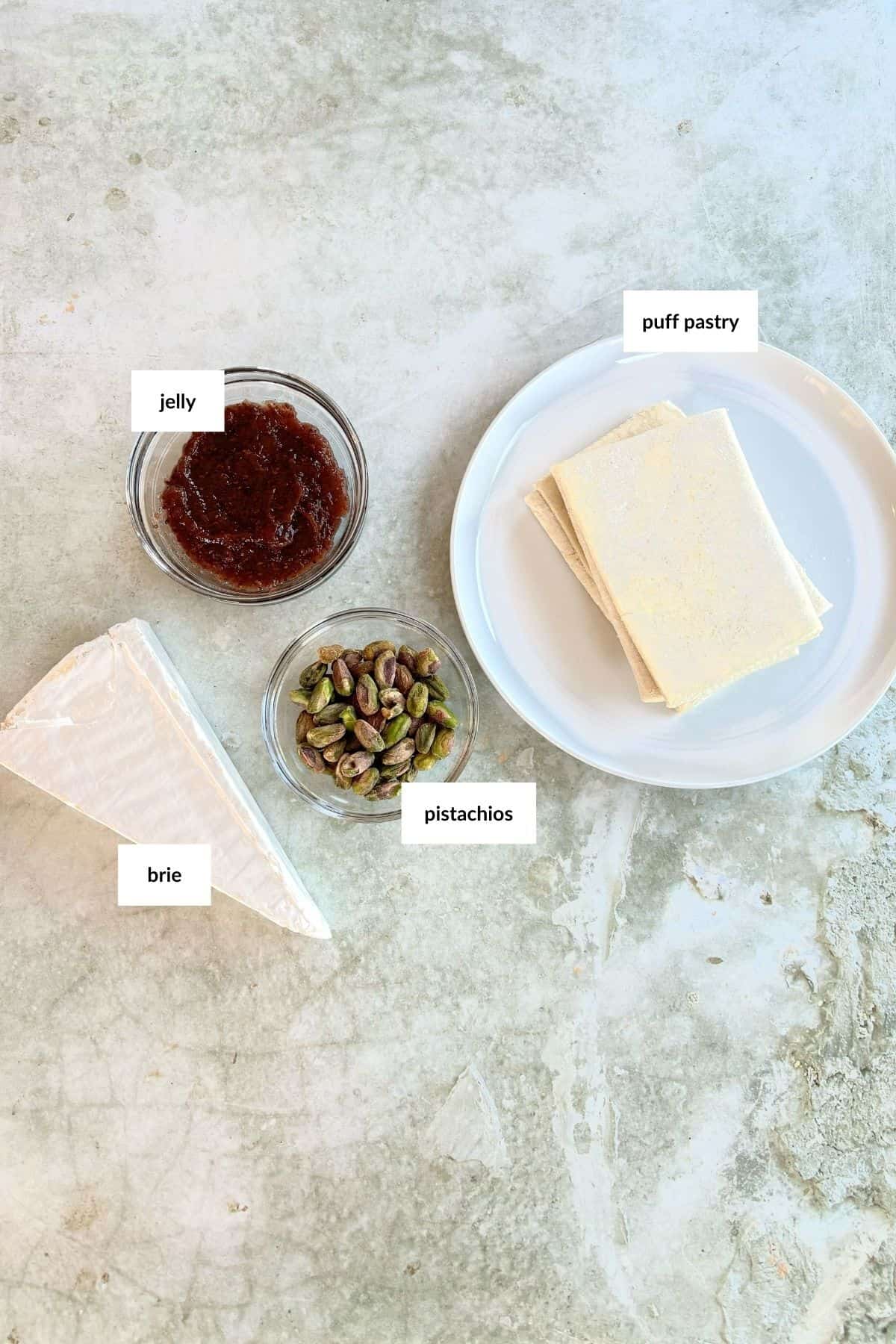 A wedge of brie, a bowl of jelly, a bowl of pistachios, and a plate with puff pastry squares on a light textured surface.