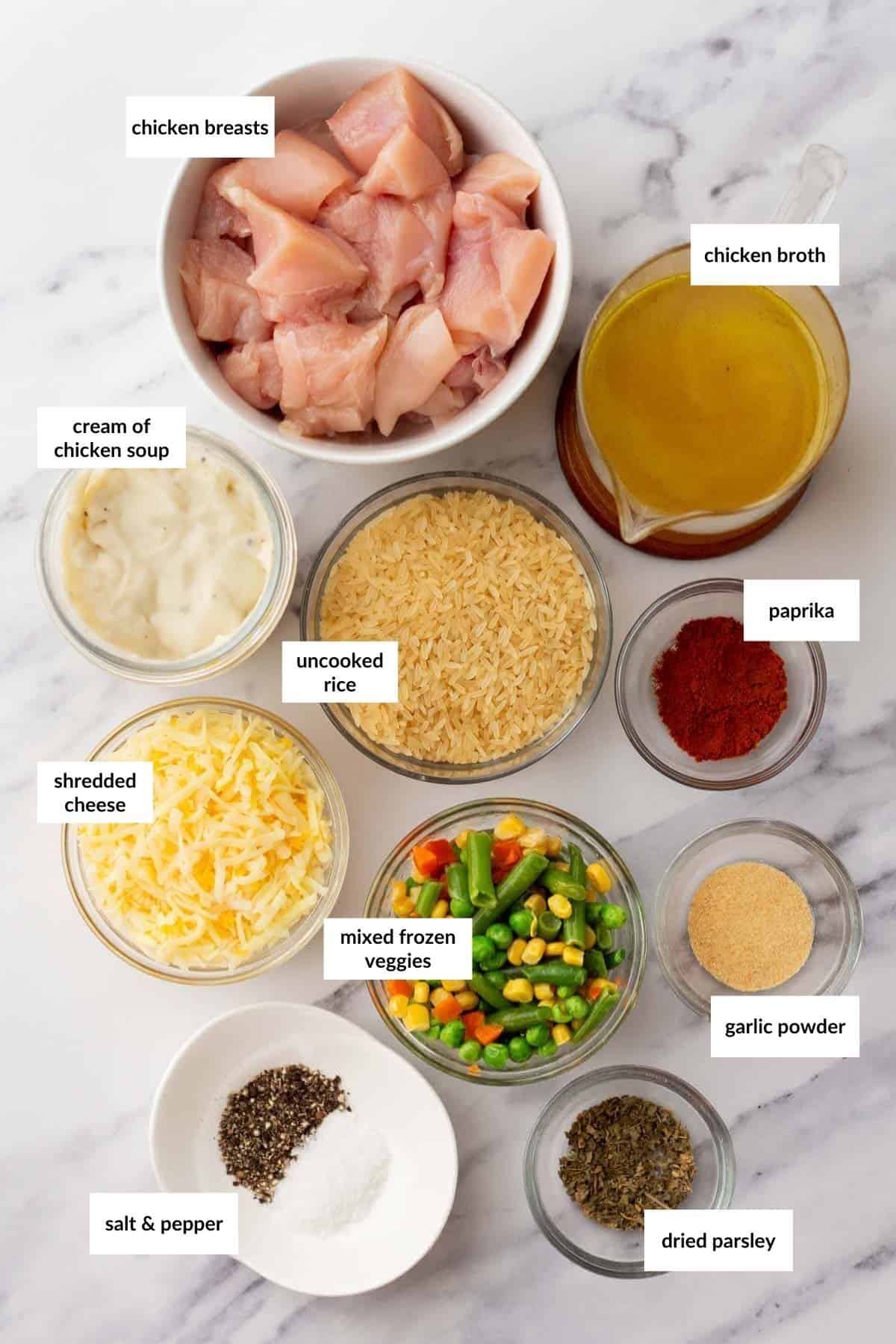 Assorted ingredients on a marble surface, including chicken breasts, chicken broth, cream of chicken soup, uncooked rice, paprika, shredded cheese, mixed frozen veggies, garlic powder, salt, and pepper.
