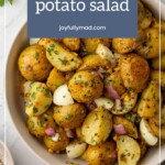 A bowl of French potato salad with halved potatoes, herbs, and chopped onions, garnished with parsley. The text overlay reads "French potato salad" followed by "joyfullymad.com". Perfect alongside chocolate buttercream frosting for a sweet and savory delight!.