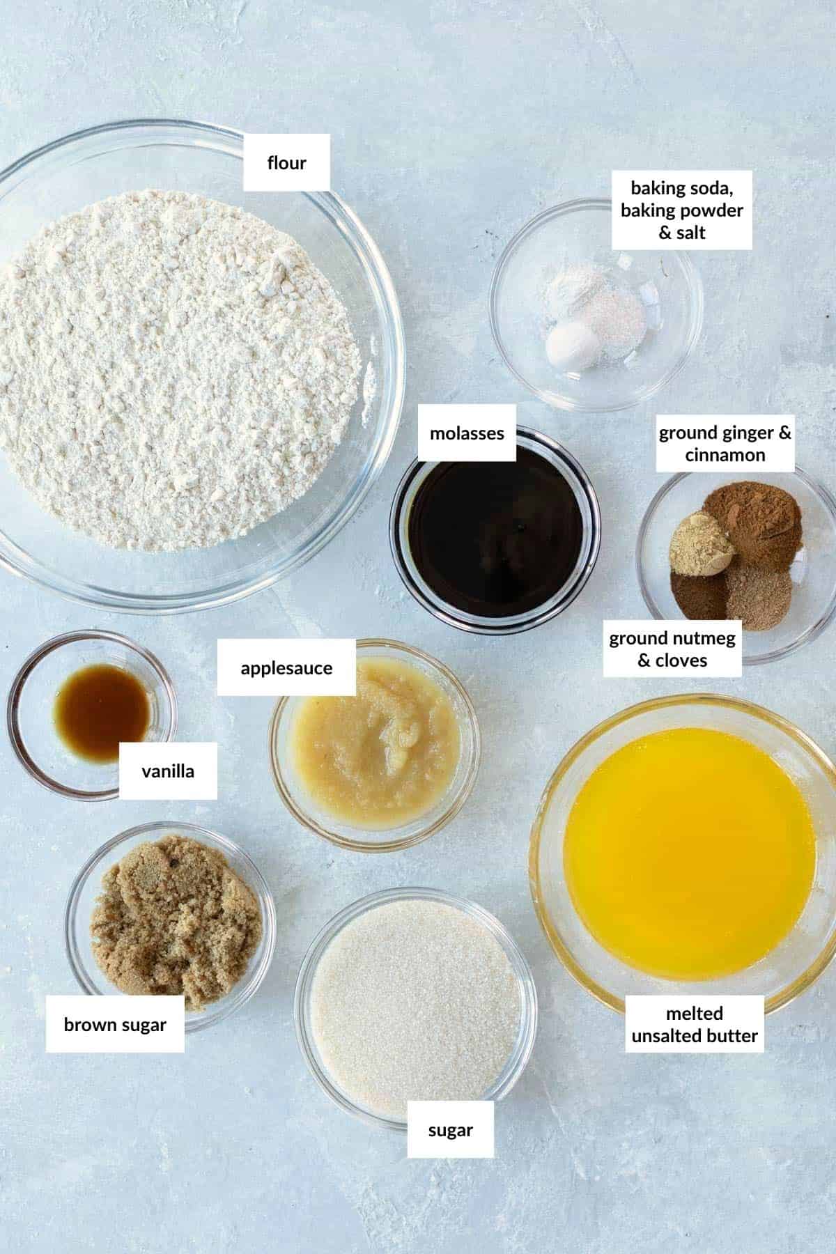 Various baking ingredients including flour, molasses, spices, sugars, melted butter, applesauce, and vanilla are arranged in labeled bowls on a light surface.