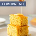 Two square pieces of cornbread stacked on a plate with honey being drizzled from above, all complemented by a rich layer of chocolate buttercream frosting.