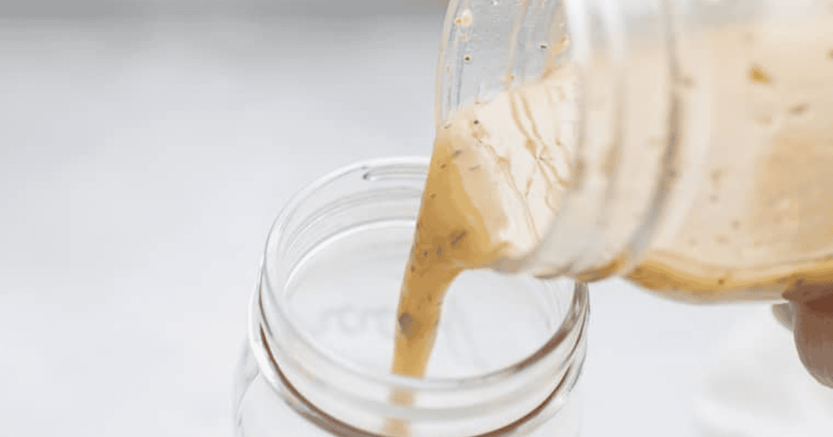 Pouring creamy dressing from a glass jar into another glass jar against a light background.