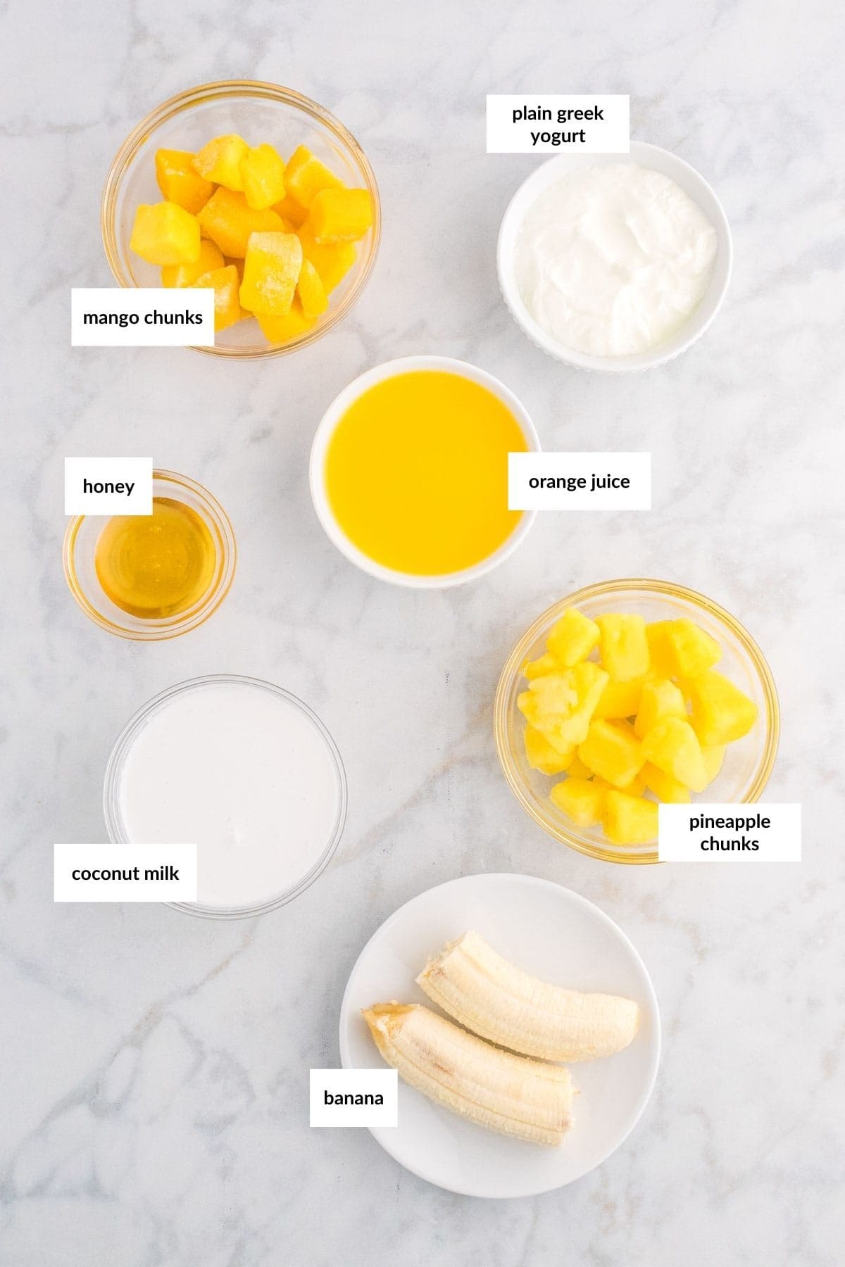 Ingredients for a smoothie arranged on a marble surface: mango chunks, honey, coconut milk, banana, pineapple chunks, orange juice, and plain Greek yogurt.