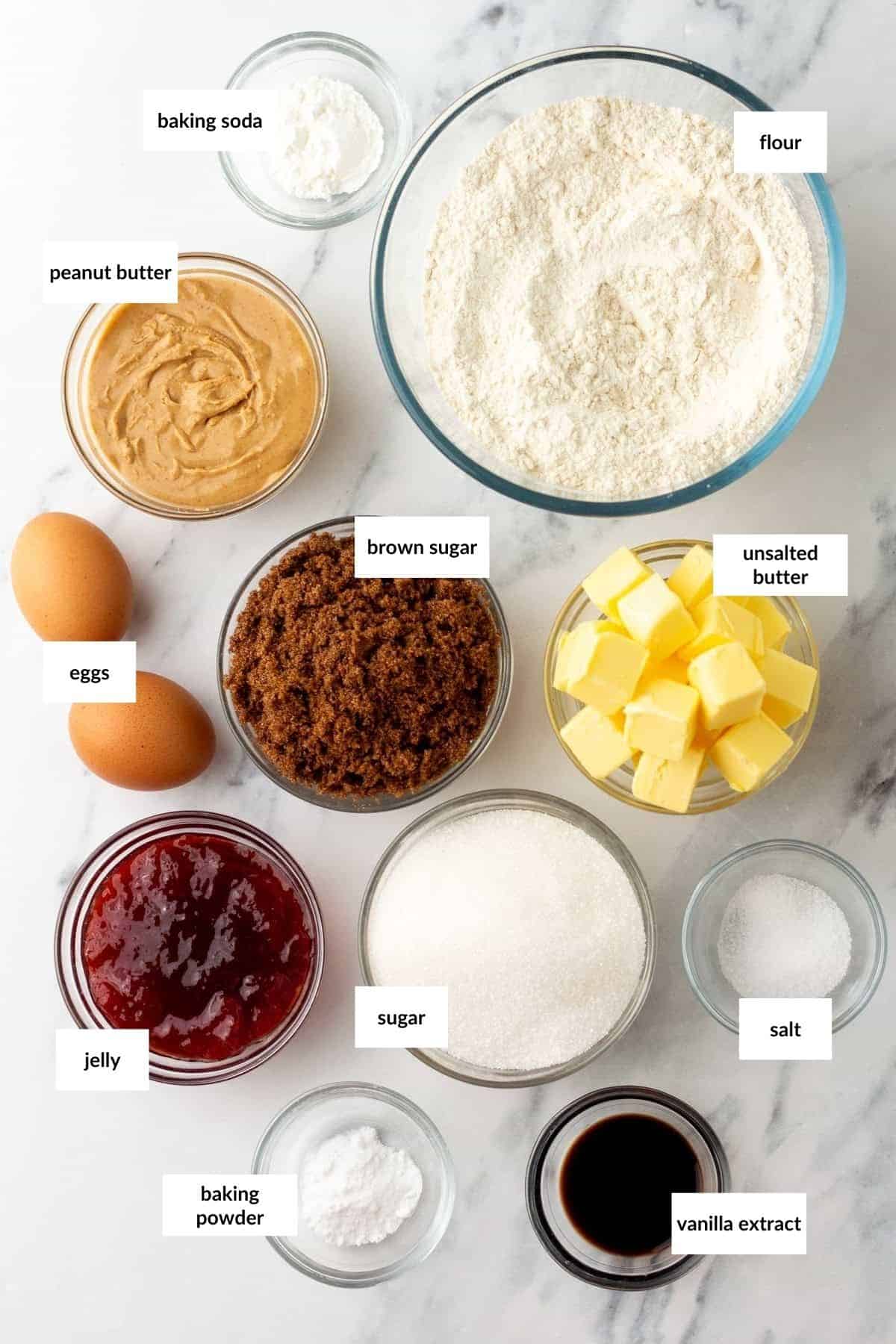 Ingredients for a baking recipe arranged on a marble surface, including flour, peanut butter, brown sugar, butter, jelly, eggs, sugar, vanilla extract, baking soda, baking powder, and salt.