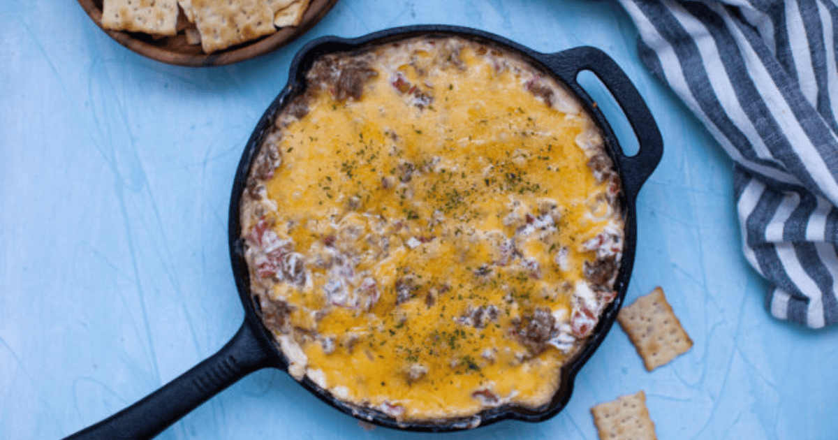 A skillet with melted cheese dip containing bits of tomato and herbs on a light blue surface, accompanied by scattered crackers and a striped cloth.