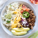 A bowl of rice, seasoned ground beef, chopped onions, grated cheese, sliced jalapeño, avocado, tomato salsa, and a lime wedge, garnished with cilantro.