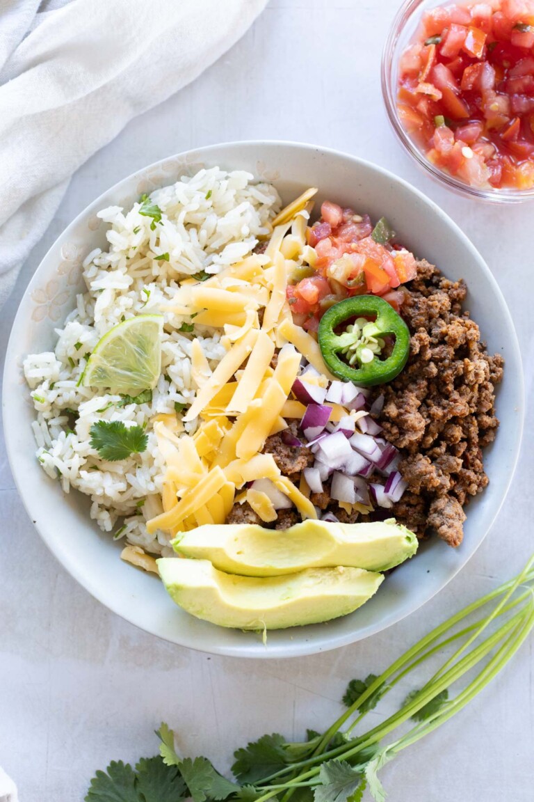 A bowl of rice, seasoned ground beef, chopped onions, grated cheese, sliced jalapeño, avocado, tomato salsa, and a lime wedge, garnished with cilantro.