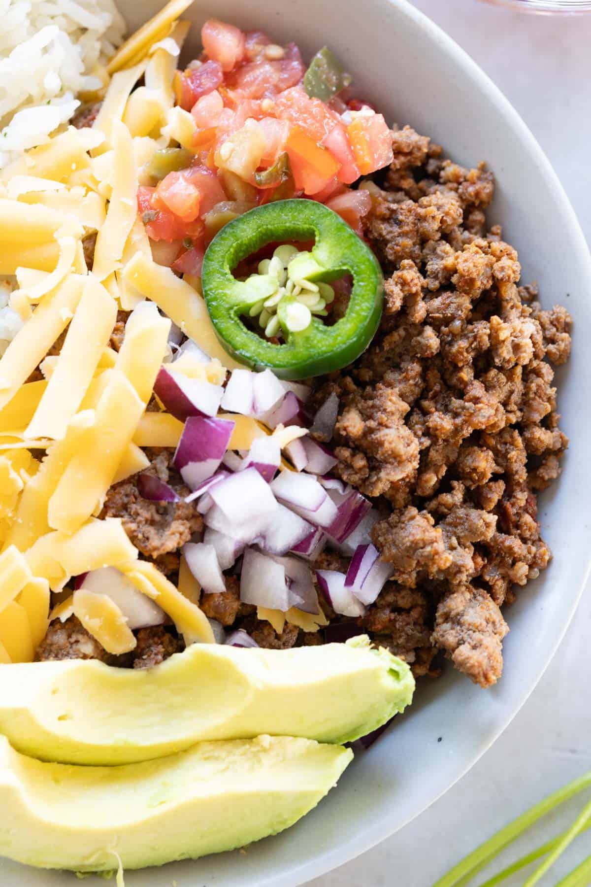 A bowl with ground beef, shredded cheese, diced onions, avocado slices, jalape&ntilde;o, tomato salsa, and rice.