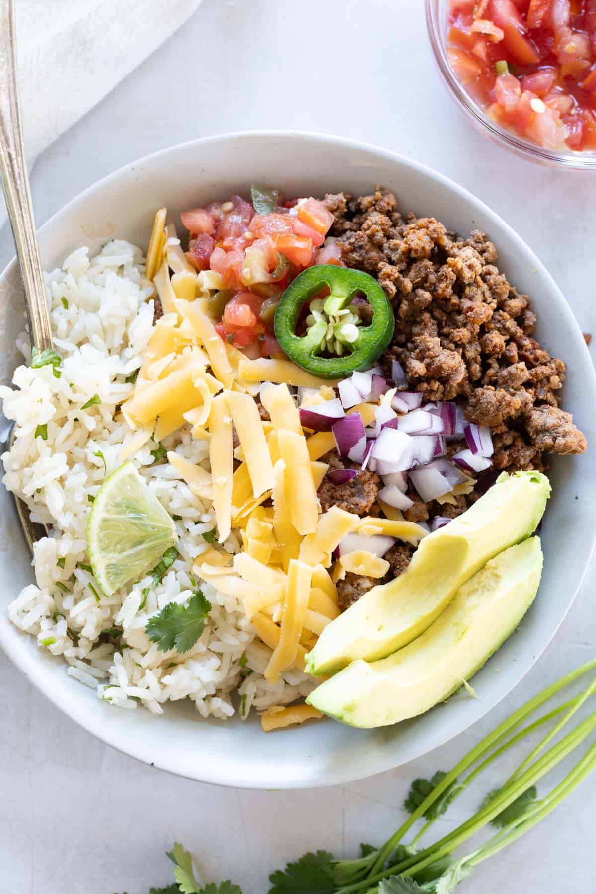 A bowl with rice, seasoned ground beef, avocado slices, shredded cheese, diced red onion, jalape&ntilde;o, lime, pico de gallo, and cilantro.