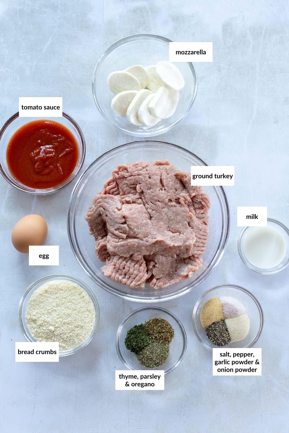 Ingredients for a recipe including ground turkey, mozzarella, tomato sauce, egg, bread crumbs, herbs, spices, and milk arranged in bowls on a light surface.
