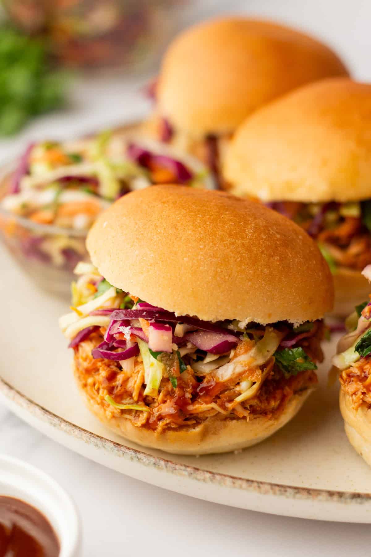 Pulled chicken sliders topped with coleslaw on a platter, with a small bowl of sauce in the foreground.