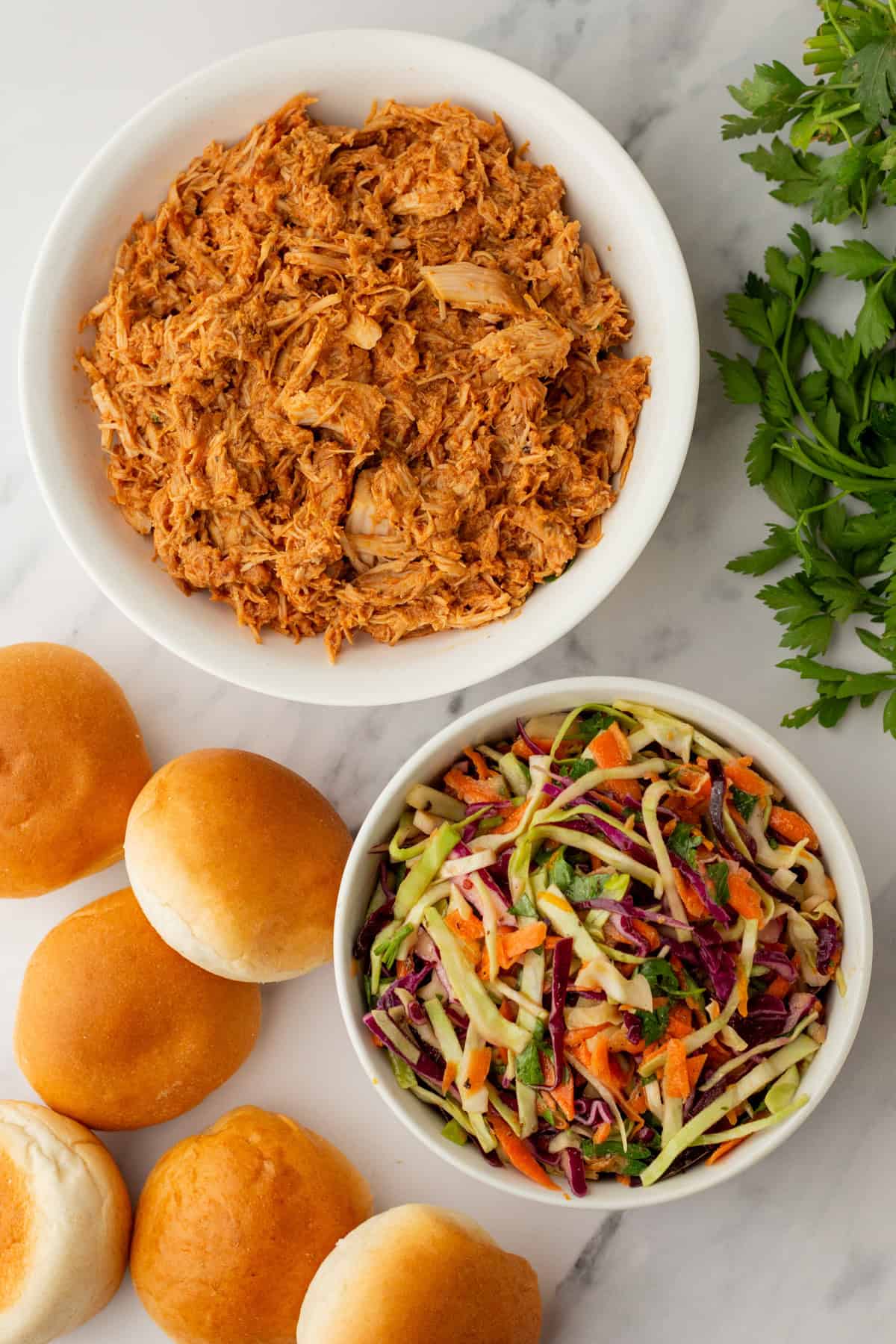 Shredded chicken in a bowl, coleslaw in another bowl, four bread rolls, and fresh parsley on a marble surface.
