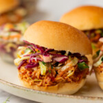 A food plate with three pulled pork sliders topped with coleslaw in soft buns, garnished with fresh greens in the foreground.