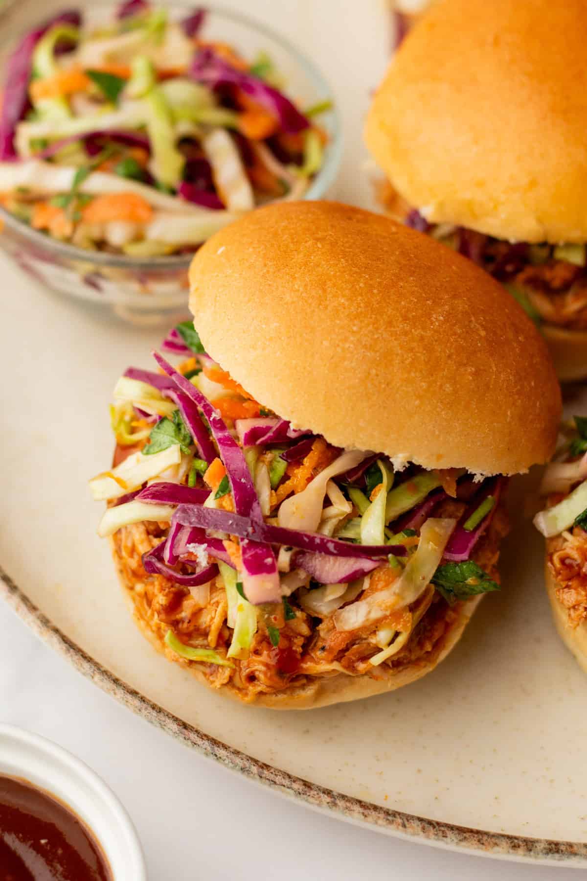 Pulled pork sandwich topped with vibrant coleslaw on a bun, accompanied by a bowl of coleslaw and a small dish of barbecue sauce on a light-colored plate.
