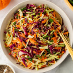 A bowl of mixed cabbage slaw with carrots and herbs, placed on a marble surface. A spoon rests in the bowl. Nearby are oranges, a bowl of pepper, and a wooden bowl with arugula.