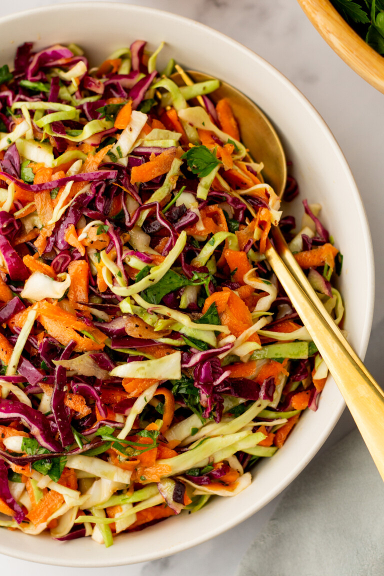 A bowl of coleslaw featuring shredded green and purple cabbage, carrots, and parsley, with a gold serving spoon on the side.