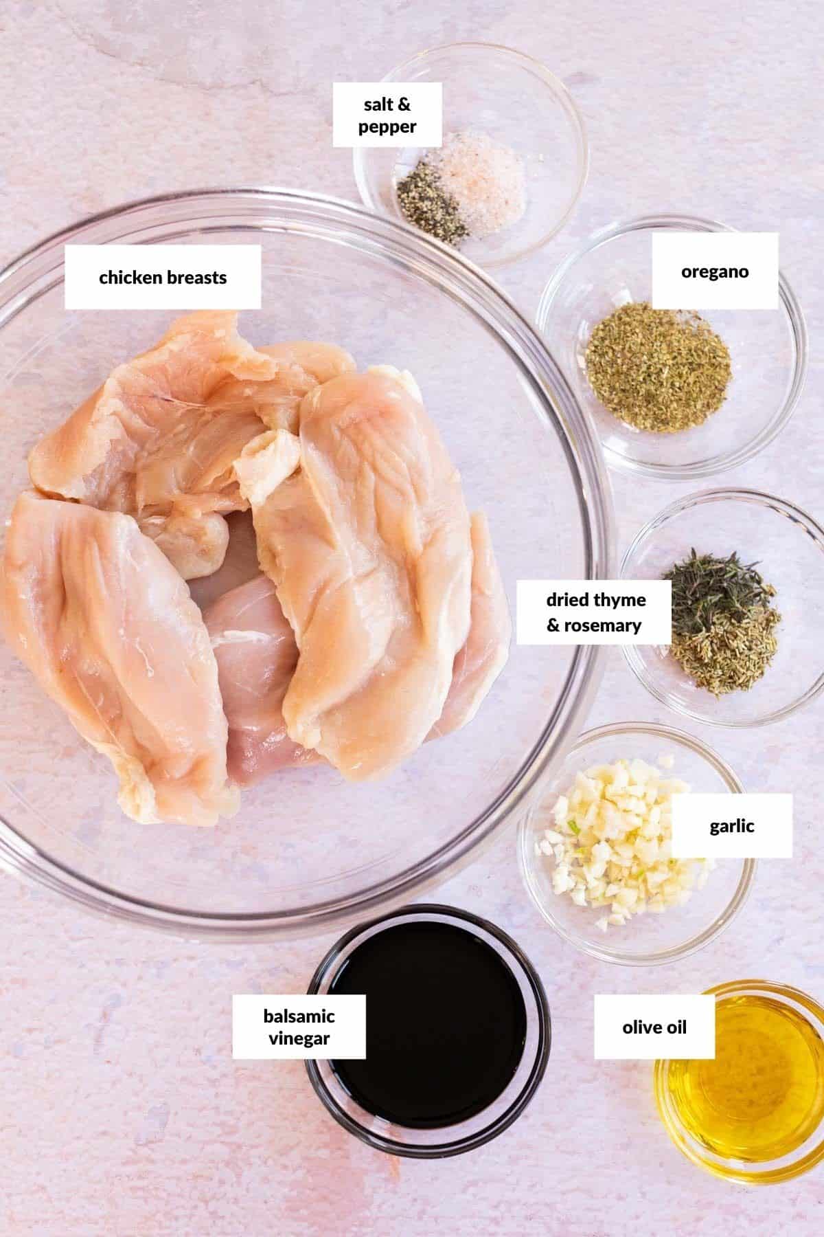 Raw chicken breasts in a bowl surrounded by small bowls of salt and pepper, oregano, dried thyme and rosemary, minced garlic, balsamic vinegar, and olive oil, all labeled.