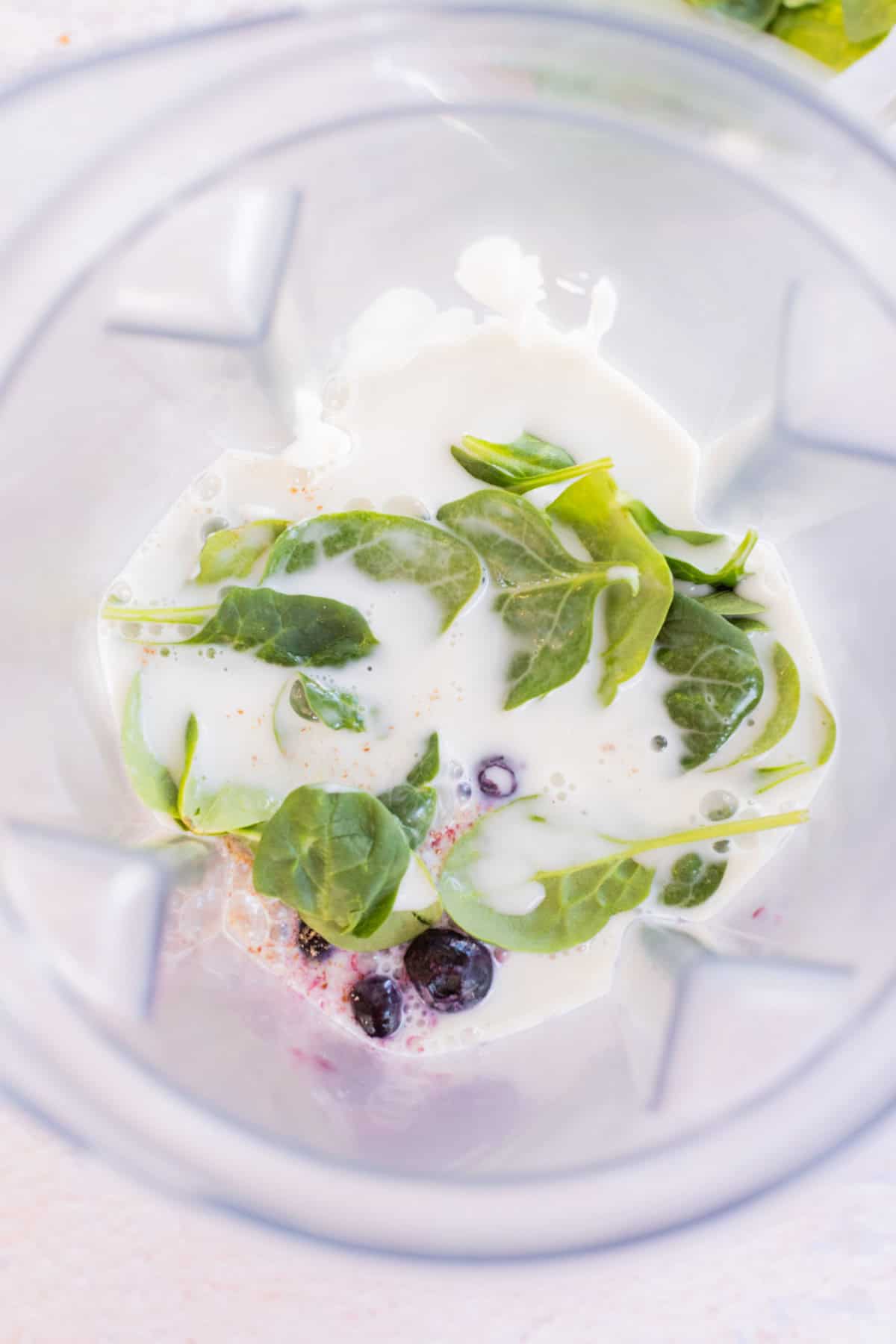 Spinach leaves, blueberries, and milk in a blender, viewed from above.