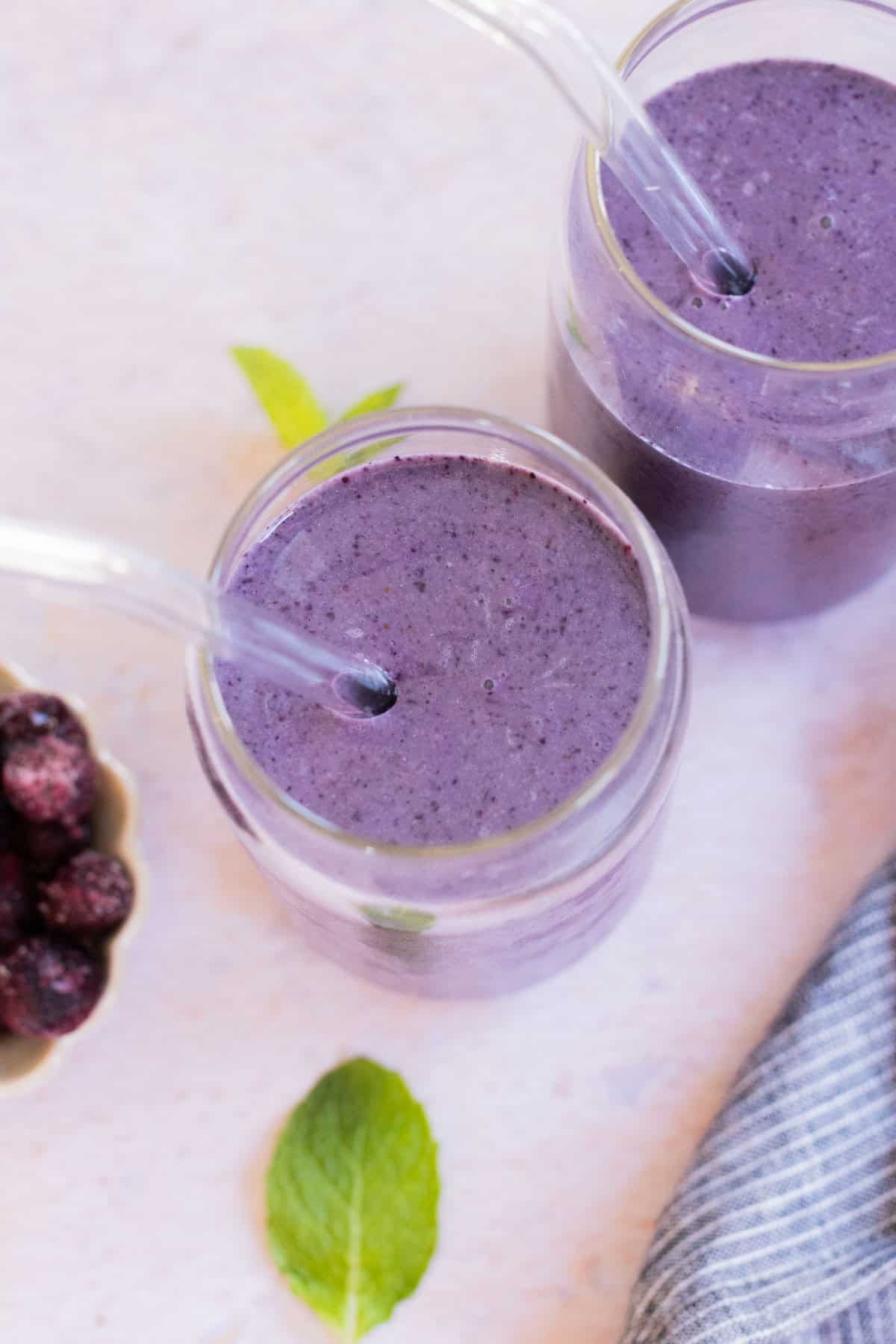 Two glass jars of blueberry smoothie with clear straws on a light surface. A bowl of berries and a mint leaf are beside them. A striped cloth is partially visible.